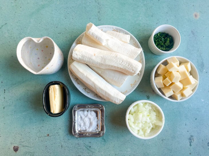 Ingredientes separados na bancada com aipim, queijo coalho, cebolas, salsinha, cebolinha e margarina organizados para o preparo.