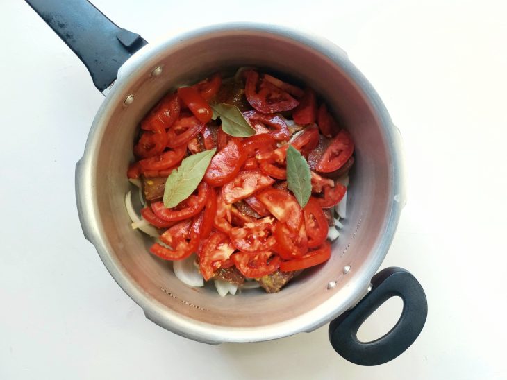 Uma panela de pressão com camadas de cebolas, de carne, tomates picados e louro.