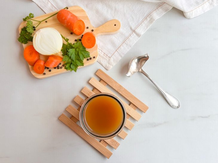 Caldo pronto em um vidro, legumes e talheres acompanham a foto.