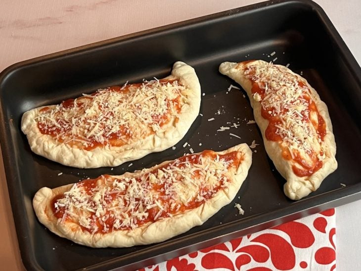 Uma forma contendo 3 calzones crus pincelados com molho de tomate e polvilhados com queijo.