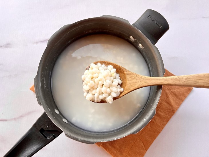 Uma colher com canjica ou mungunzá cozido retirado da panela de pressão ao fundo.