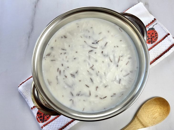 Uma panela cozinhando a canjiquinha com leite, leite condensado, leite de coco, coco ralado, canela em pau e cravos-da-índia.