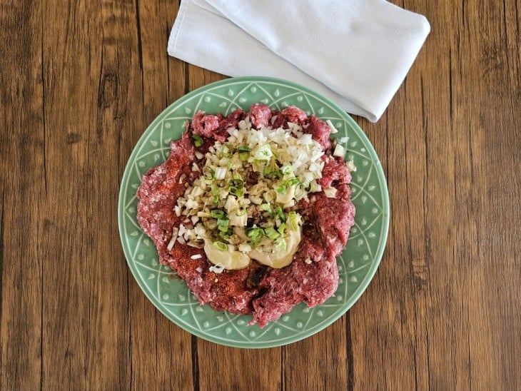 Um prato contendo carne moída, páprica, pimenta calabresa, alho, cebola, conhaque, mostarda, azeite e colheres de cebolinha.