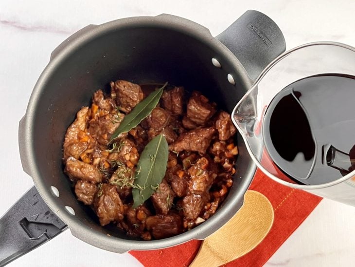 Uma panela de pressão contendo carne, molho de tomate, folhas de louro e vinho tinto.