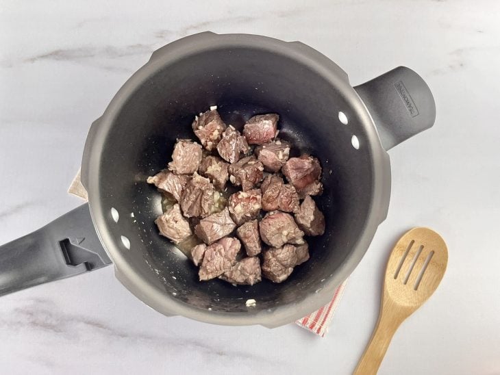 Uma panela de pressão refogando cebola, alho e cubos de carne.