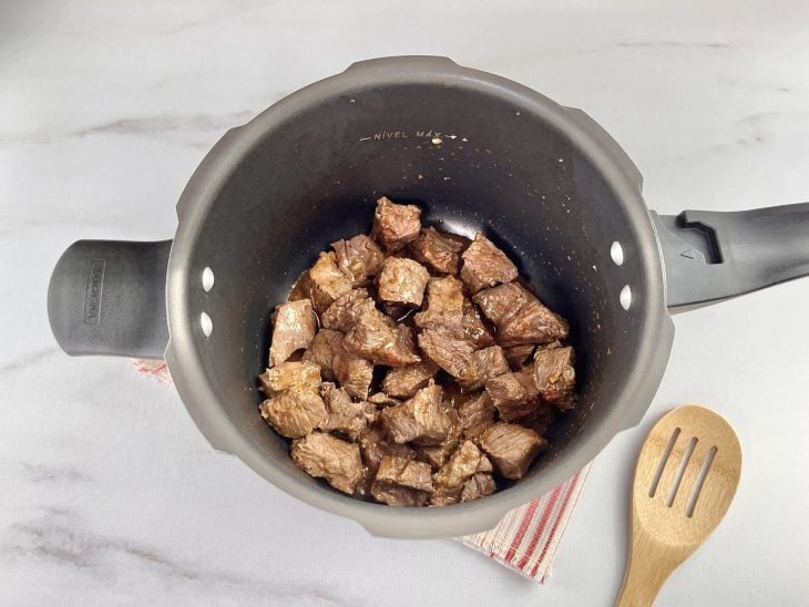 Uma panela de pressão refogando cebola, alho e cubos de carne com temperos.