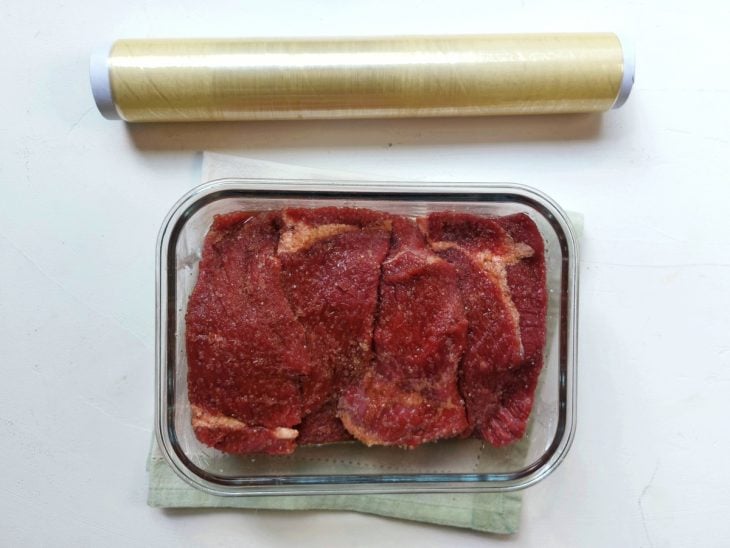 Um recipiente contendo carne com açúcar e sal, coberto com papel filme.