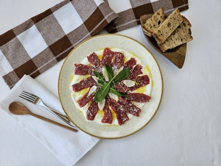 Carpaccio de carne disposto em um prato redondo com pães ao lado.