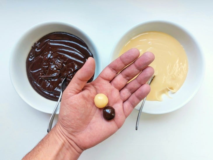 Uma bolinha de brigadeiro e outra de brigadeiro branco na mão. Com os pratos dos dois preparos ao fundo.