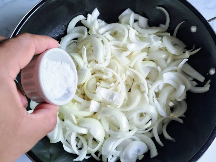 Uma frigideira refogando cebolas cortadas com sal.