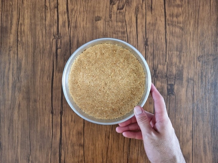 Mão segurando a forma com a massa de bolachas apertada nas laterais e ao fundo.