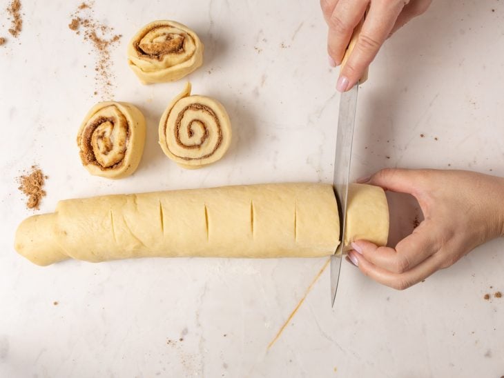 Rolo de massa de cinnamon rolls sendo fatiado para formar os pãezinhos.