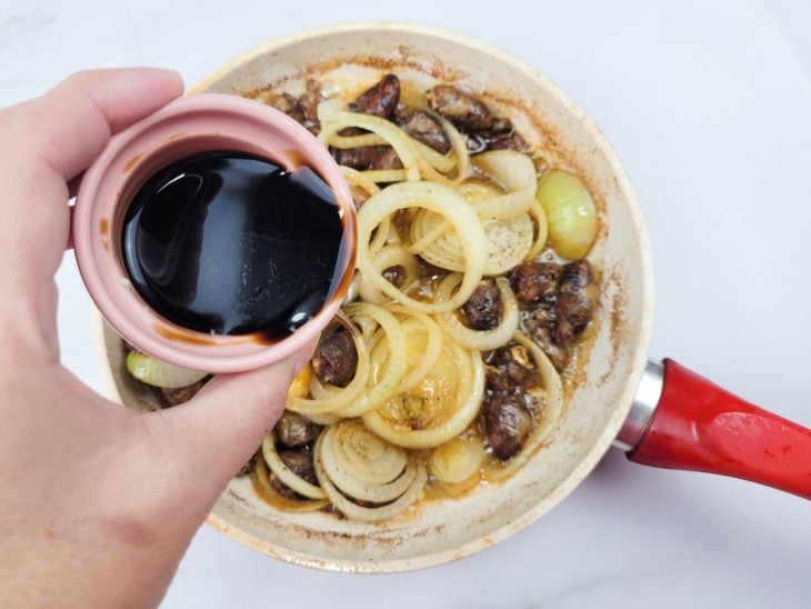 Uma frigideira refogando os corações de galinha na manteiga, cobertos com rodelas de cebola com shoyu.