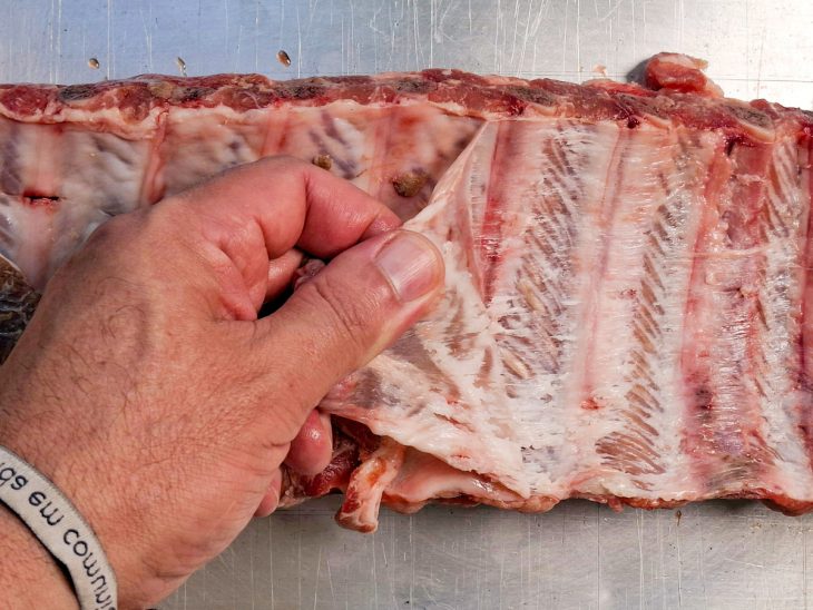 Costelinha sobre a tábua com a membrana sendo retirada com as mãos.