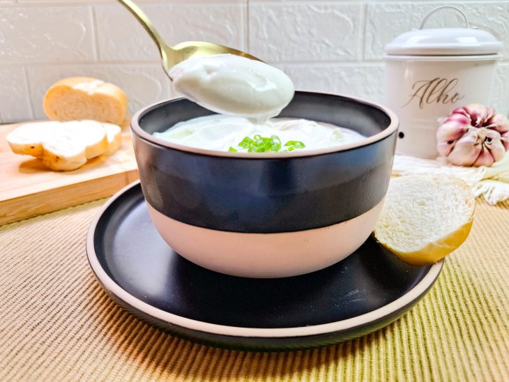 Na tigela, o creme de alho, colher tirando uma porção, fatia de pão acompanha o quadro.