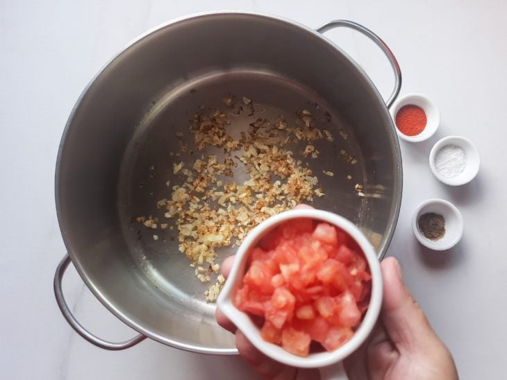 Uma panela com alho e cebola dourados e recipientes com tomate e temperos.