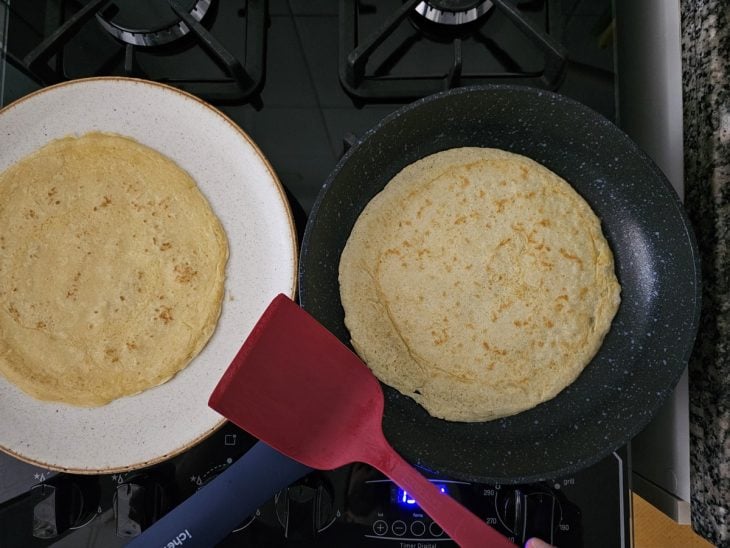 Um prato com crepe pronto e outra com a frigideira e mais crepes sendo feitos.