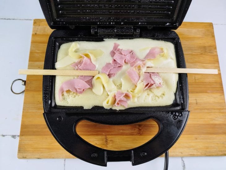 Uma sanduicheira com a massa do crepe, o recheio e palitos.