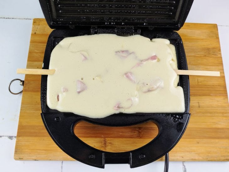 Uma sanduicheira com a massa do crepe, o recheio e palitos.