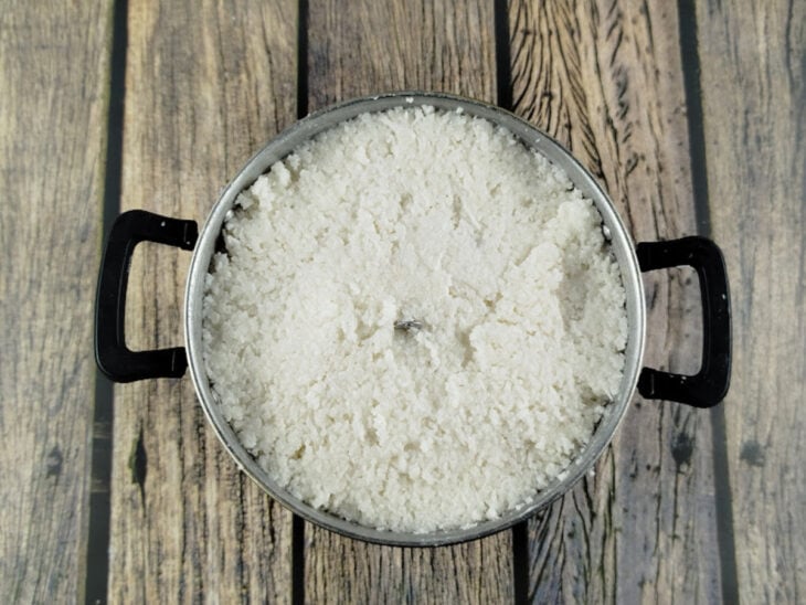 Massa de tapioca sendo colocada delicadamente e sem pressão dentro de uma cuscuzeira grande.