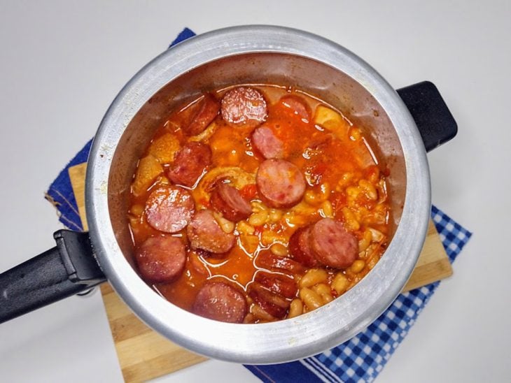 Uma panela de pressão contendo cebola, alho, tomates, bucho, feijão-branco, molho de tomate e calabresas.