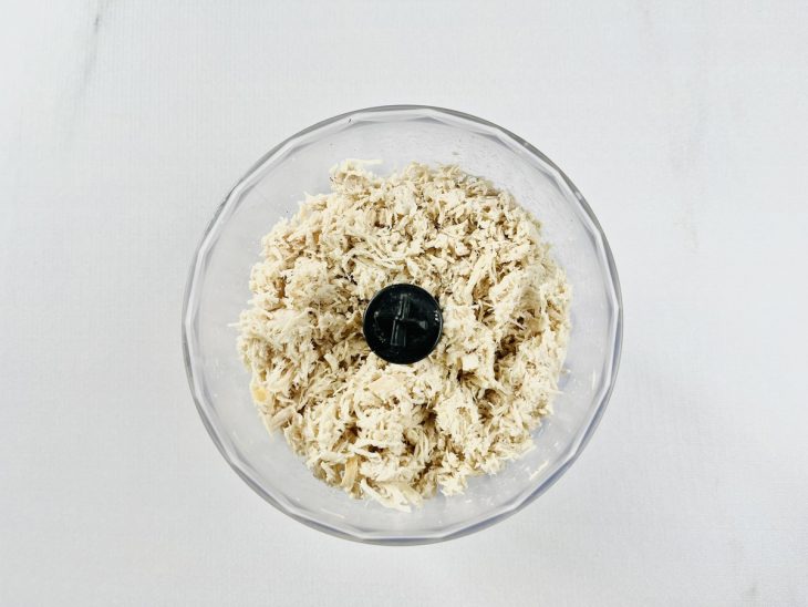 Frango cozido sendo desfiado com garfo sobre prato fundo.