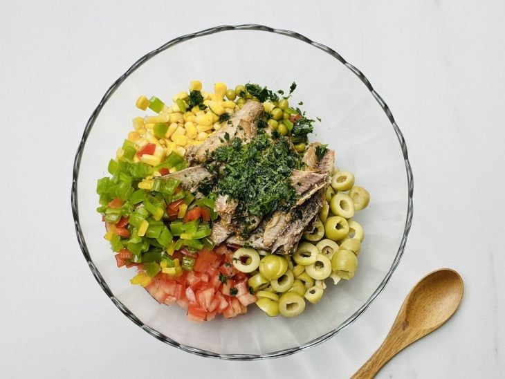Uma tigela com a mistura de tomates, pimentão, cebola, milho, ervilha, azeitonas, cheiro-verde e sardinhas.