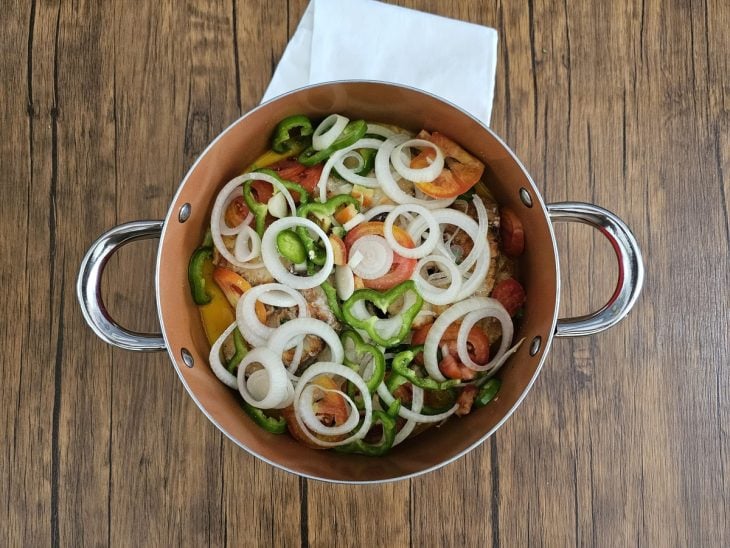 Uma panela cozinhando postas de peixes com vegetais, rodelas de cebola, rodelas de tomate e leite de coco.