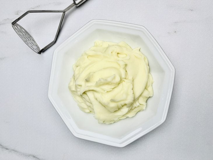 Batatas cozidas amassadas em um prato com auxílio de um amassador de batatas.