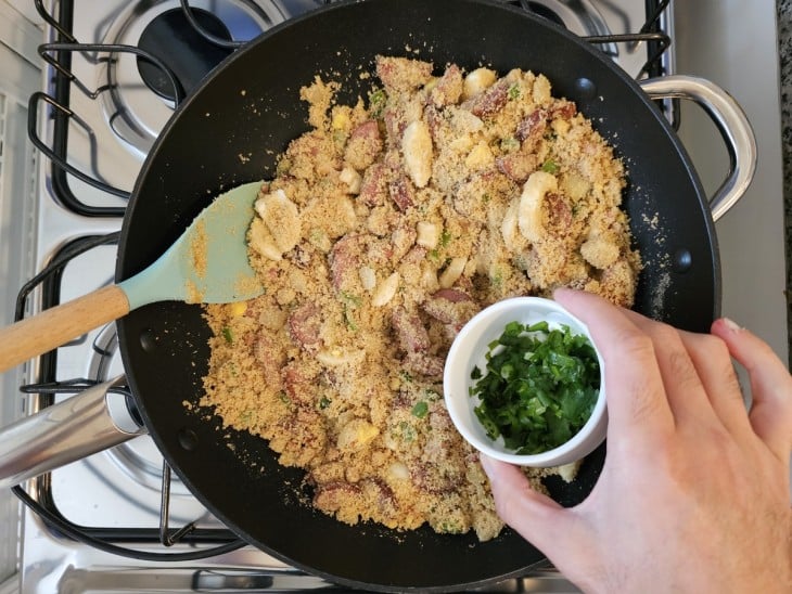 Uma frigideira com bacon, calabresa, legumes, farinha de milho refogados e cheiro-verde.