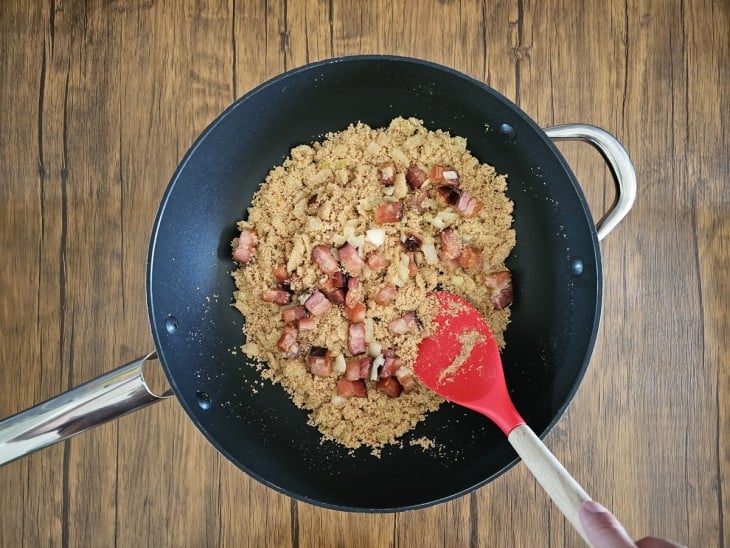 Uma frigideira com farinha de mandioca, cebola e bacon sendo refogada.