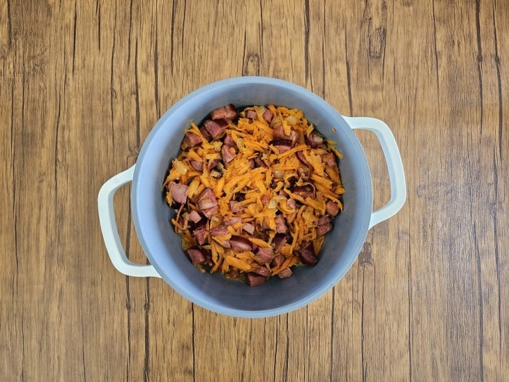 Cenoura, bacon, calabresa e cebola misturados em uma panela cinza com alças na cor branca.