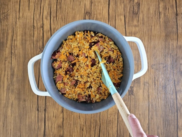 Cenoura, bacon, calabresa, cebola e farinha misturados em uma panela cinza com alças na cor branca.