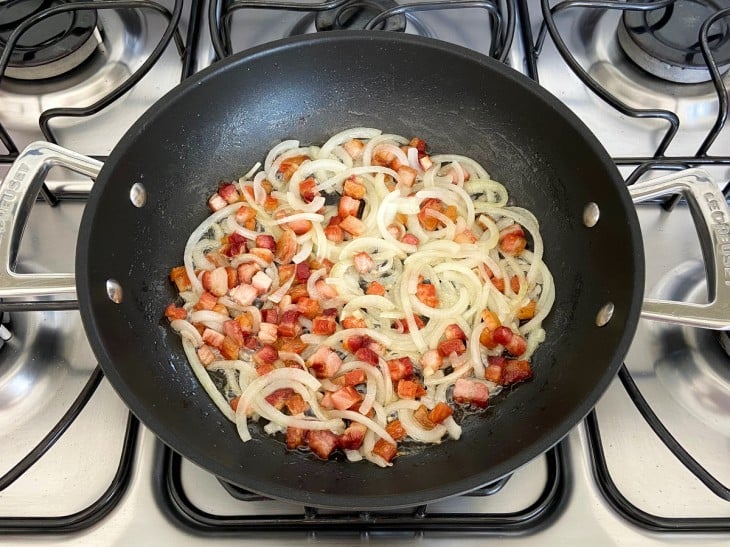 Uma frigideira com bacon em cubinhos fritos e rodelas de cebola.