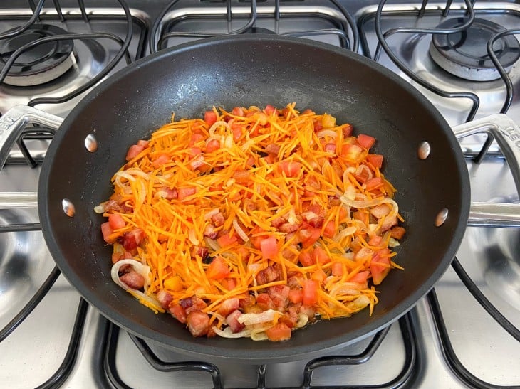 Uma frigideira com bacon em cubinhos fritos, rodelas de cebola e cenoura ralada.