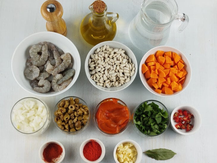 Ingredientes como feijão branco de molho, camarões limpos, temperos e azeites dispostos em potes na bancada.