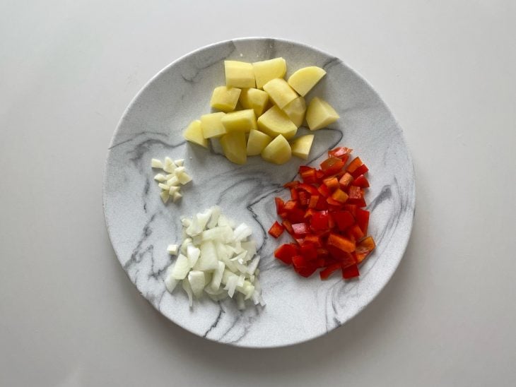 Alho, cebola, pimentão e batata sendo cortados em pedaços pequenos.
