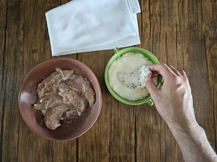 Uma vasilha com filés, outra com queijo e tapioca com um filé sendo empanado.