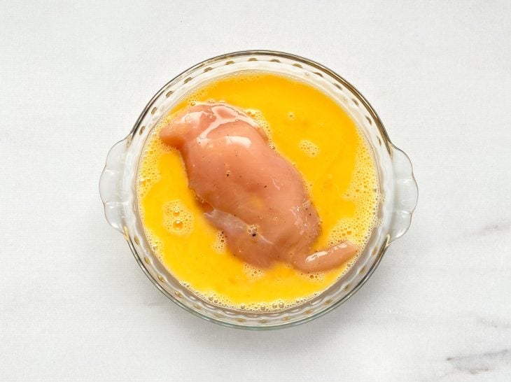 Um recipiente contendo ovos batidos empanando um filé de frango recheado.