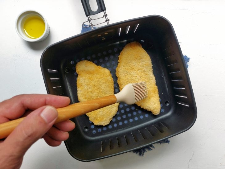 Uma cesta de airfryer contendo dois filés de peixe empanados e sendo pincelados com azeite.