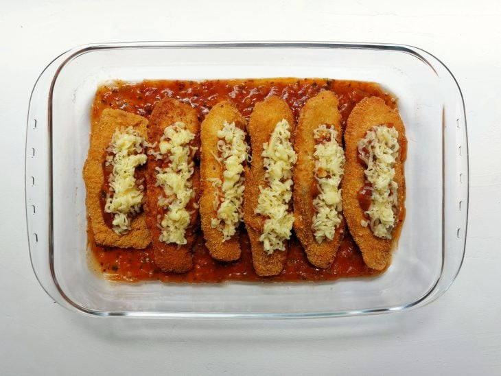 Um refratário contendo filé de tilápia empanados e fritos, cobertos com molho e queijo.