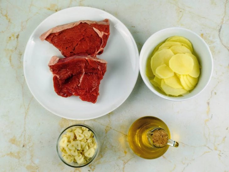 Ingredientes organizados na bancada com alho descascado e batata sendo fatiada fina.