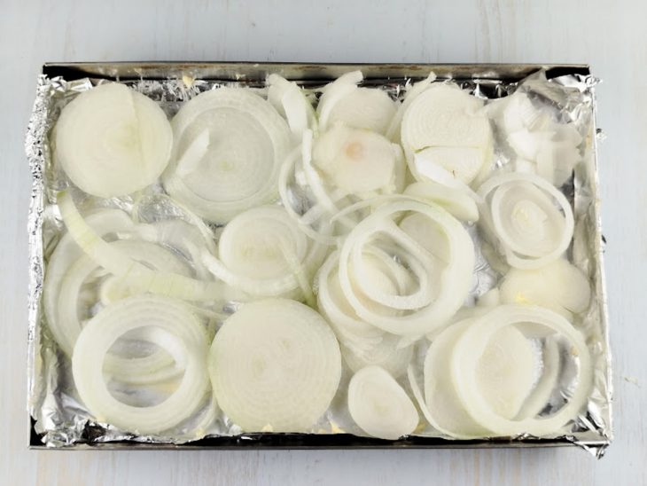 Rodelas de cebola com sal e água espalhadas sobre uma forma de alumínio.