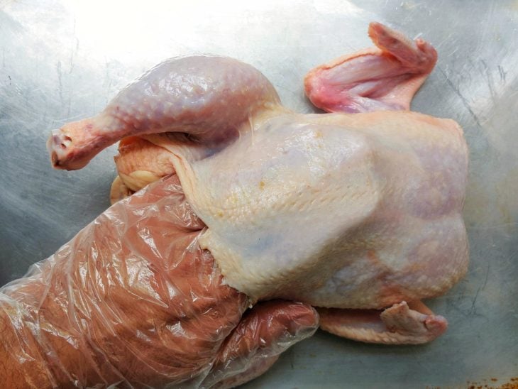 Mão com luva dentro do espaço entre a carne e a pele do frango inteiro.