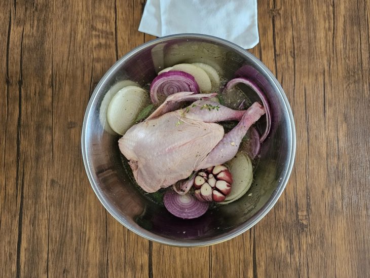 Um recipiente contendo frango galeto, rodelas de cebola e uma cabeça de alho.
