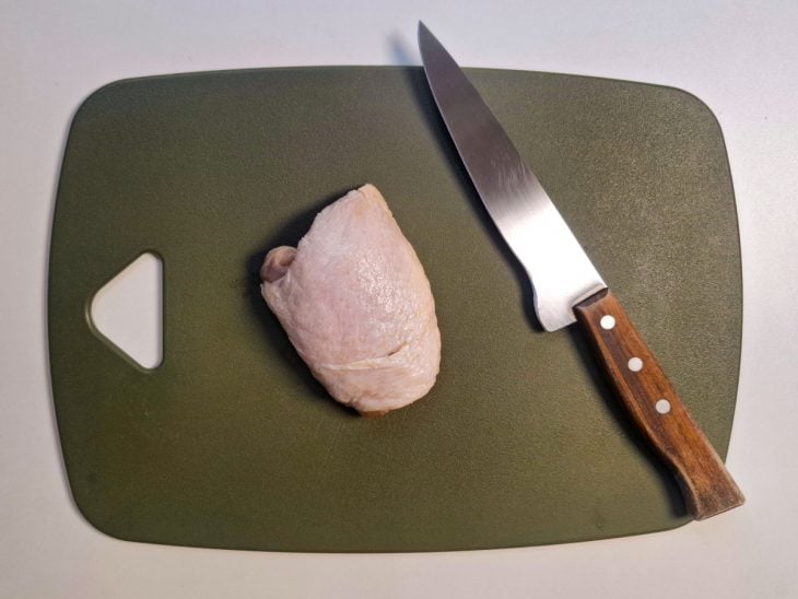 Pedaço de frango sobre uma tábua sem pedaços de gordura, já retirados pela faca posicionada ao lado.