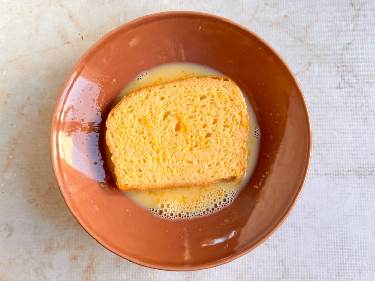 Fatia de pão brioche sendo mergulhada na mistura líquida.