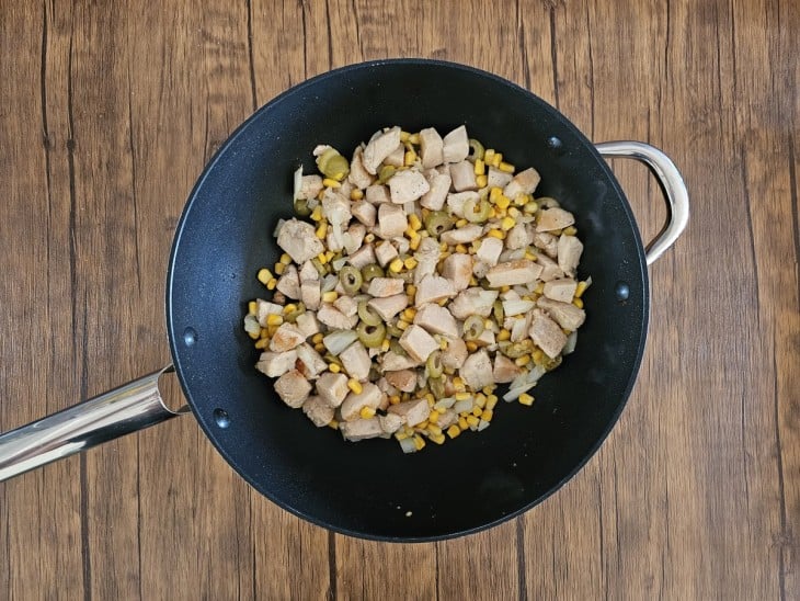 Um frigideira com cubos de frango, cebola, alho, milho e azeitonas refogados.