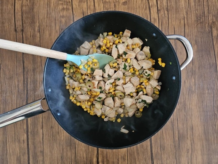 Um frigideira com cubos de frango, cebola, alho, milho e azeitonas refogados.