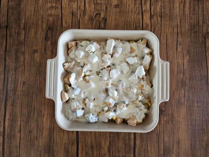 Uma forma com cubos de frango, cebola, alho, milho e azeitonas refogados cobertos por molho.